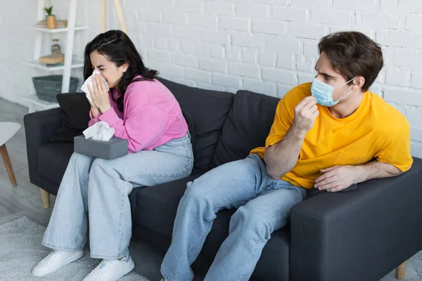 Diseased young woman sitting on couch and blowing nose with paper napkin near boyfriend adjusting medical mask at home — Stock Photo