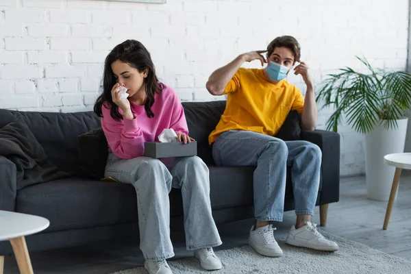Diseased young woman blowing nose with paper napkin near boyfriend sitting on couch and adjusting medical mask at home — Stock Photo