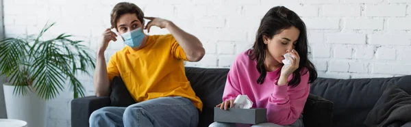 Diseased young woman blowing nose near boyfriend sitting on couch and adjusting medical mask at home, banner — Stock Photo