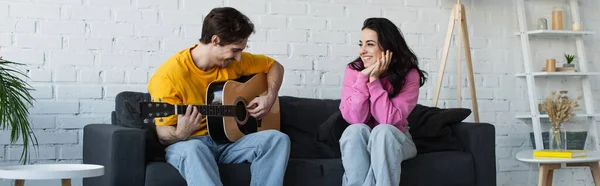 Giovane che suona la chitarra acustica vicino alla ragazza con le mani vicino al viso a casa, striscione — Stock Photo