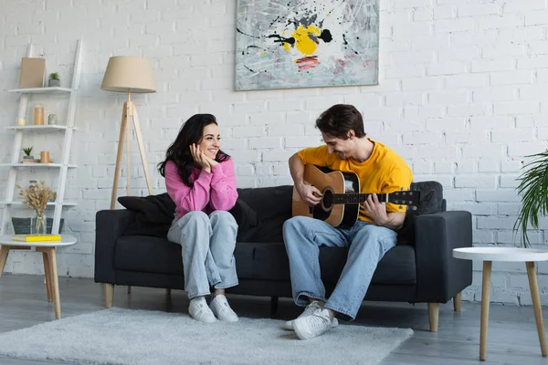 Молодий чоловік грає на акустичній гітарі біля дівчини з руками біля обличчя вдома — стокове фото