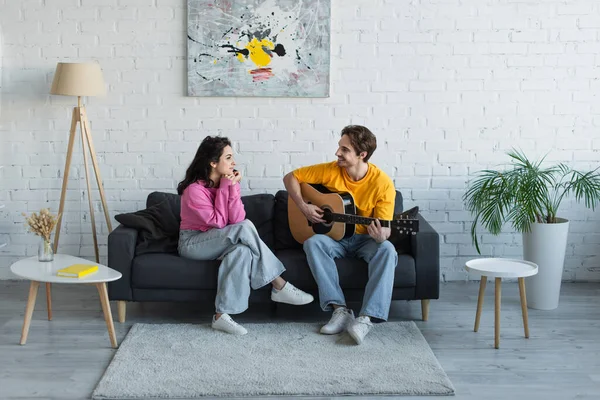 Jeune homme souriant assis sur le canapé et jouant de la guitare acoustique près de la petite amie avec les mains près du visage à la maison — Photo de stock