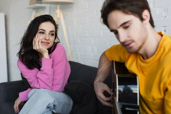 Sorridente giovane donna seduta sul divano con la mano vicino al viso vicino fidanzato offuscata suonare la chitarra acustica a casa — Foto stock