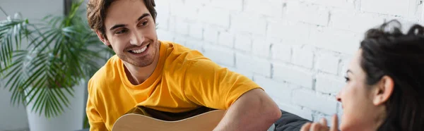 Lächelnder junger Mann sitzt auf Couch mit Akustikgitarre neben Freundin zu Hause, Transparent — Stockfoto