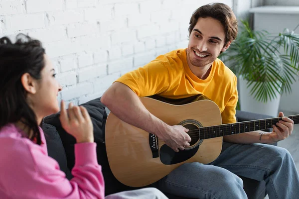 Sorridente giovane seduto sul divano con chitarra acustica vicino alla ragazza a casa — Foto stock