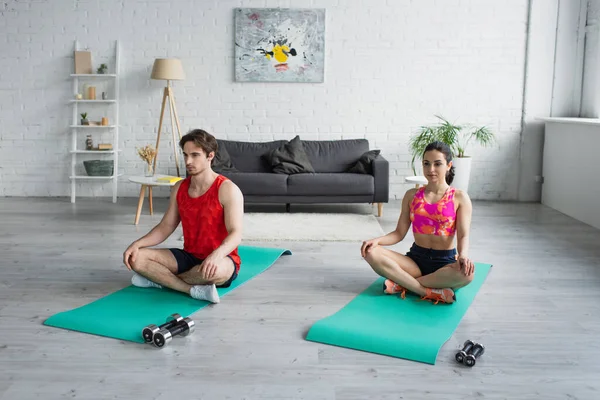 Pareja joven en ropa deportiva sentada en colchonetas de fitness en postura de yoga en casa - foto de stock
