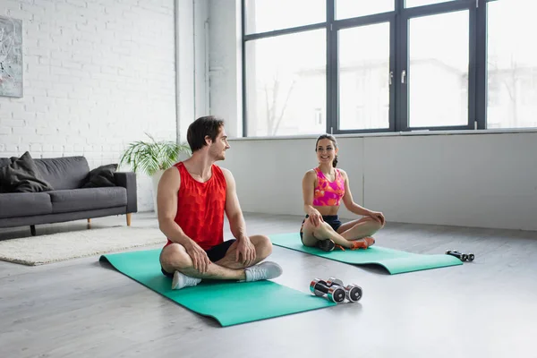 Lächelndes junges Paar in Sportbekleidung sitzt zu Hause mit überkreuzten Beinen auf Fitnessmatten — Stockfoto