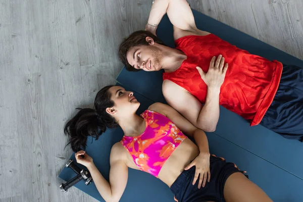 Vista superior de la joven pareja en ropa deportiva acostada en colchonetas de fitness cerca de pesas y mirándose en casa - foto de stock