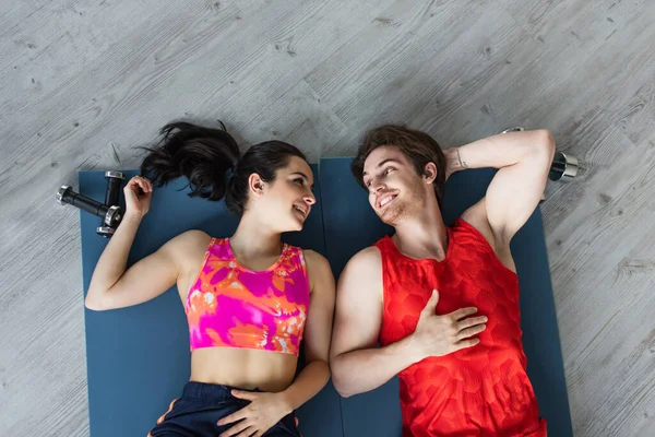 Vista superior de la joven pareja sonriente acostada en colchonetas de fitness cerca de pesas y mirándose en casa - foto de stock