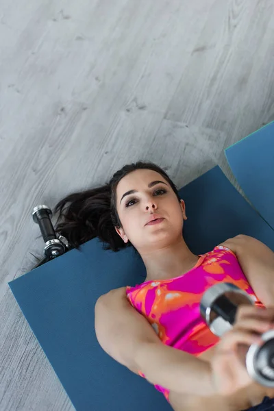 Vista superior de la joven enfocada en ropa deportiva acostada en alfombras de fitness y haciendo ejercicio con pesas en casa - foto de stock