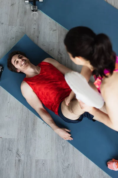 Smiling young man lying on fitness mat with outstretched leg in hands of blurred woman at home — Stock Photo