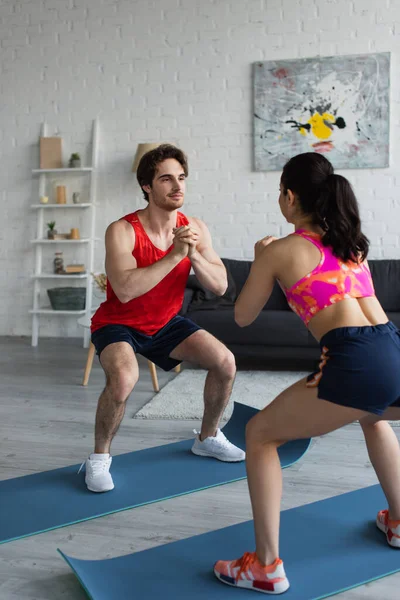 Sonriente joven pareja en ropa deportiva haciendo sentadillas en colchonetas de fitness en loft moderno - foto de stock