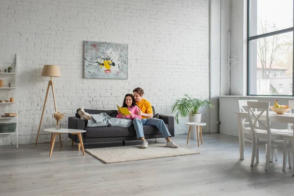 Sourire jeune homme étreignant petite amie couché sur le canapé avec livre dans les mains dans le salon — Photo de stock