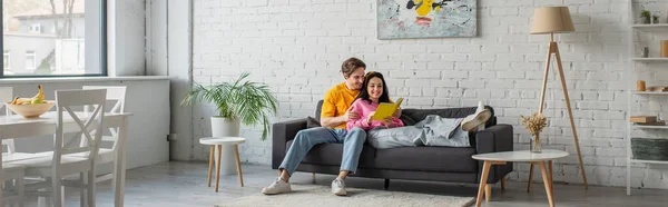 Sonriente joven abrazando novia acostado en el sofá con libro en las manos en la sala de estar, pancarta - foto de stock