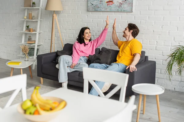 Brincalhão jovem casal sentado no sofá com alto cinco gesto na sala de estar — Fotografia de Stock