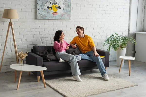 Lächelndes junges Paar, das auf der Couch sitzt und sich im modernen Wohnzimmer anschaut — Stockfoto