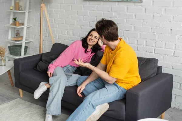 Lächelnde junge Frau sitzt auf Couch neben Freund und gestikuliert mit ausgestreckter Hand im Wohnzimmer — Stockfoto