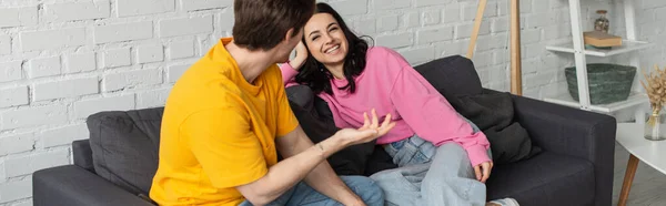 Smiling young woman sitting on couch near boyfriend gesturing with outstretched hand in living room, banner — Stock Photo