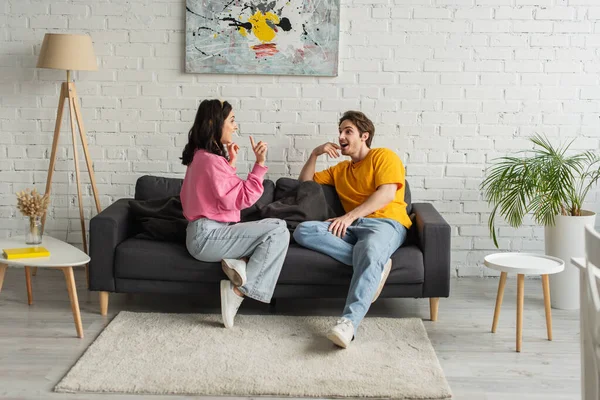 Lächelndes junges Paar sitzt auf Couch und gestikuliert mit den Händen im Wohnzimmer — Stockfoto