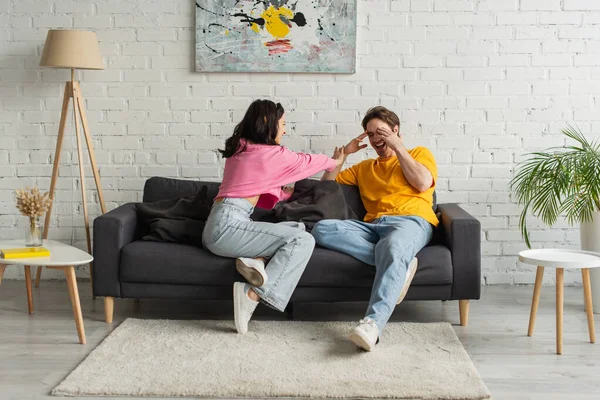 Juguetona mujer joven tocando hombre cubriendo la cara con las manos en el sofá en la sala de estar - foto de stock