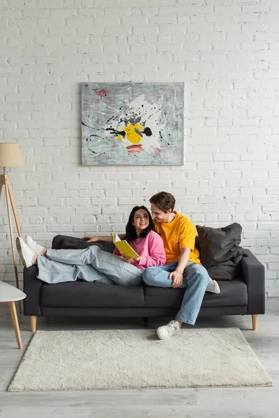 Feliz joven abrazando a la mujer con el libro acostado en el sofá en la sala de estar - foto de stock