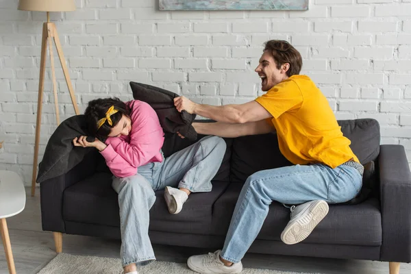 Playful young couple fighting with pillows on couch in living room — Stock Photo