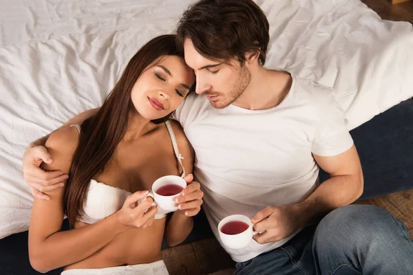 Hombre en camiseta abrazando novia seductora mientras bebe té en el dormitorio - foto de stock