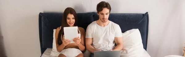 Mujer sensual en ropa interior y hombre en camiseta descansando con gadgets en el dormitorio, pancarta - foto de stock