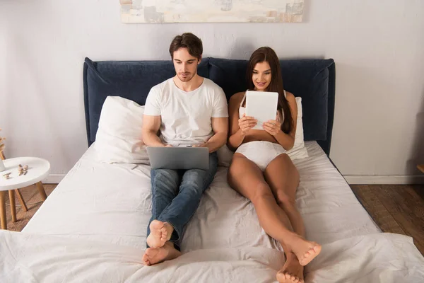 Uomo in jeans con fidanzata in lingerie sexy utilizzando dispositivi a letto — Foto stock