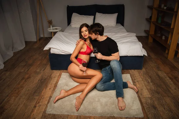 Man in jeans embracing sexy woman in red lingerie while sitting on floor with wine — Stock Photo