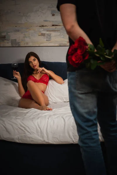 Rückansicht eines Mannes mit Rosen neben einer Frau in roten Dessous, die mit Rotwein liegt — Stockfoto