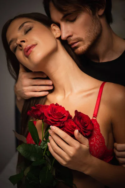 Sexy mujer con rosas rojas cerca de hombre joven abrazándola por la noche - foto de stock