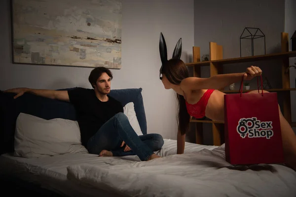 Femme sexy dans les oreilles de lapin et lingerie rouge tenant sac à provisions avec lettrage sex shop près du petit ami dans la chambre — Photo de stock