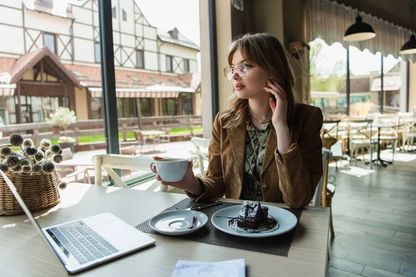 Femme à la mode dans les lunettes tenant tasse près de dessert savoureux et ordinateur portable dans le café — Photo de stock
