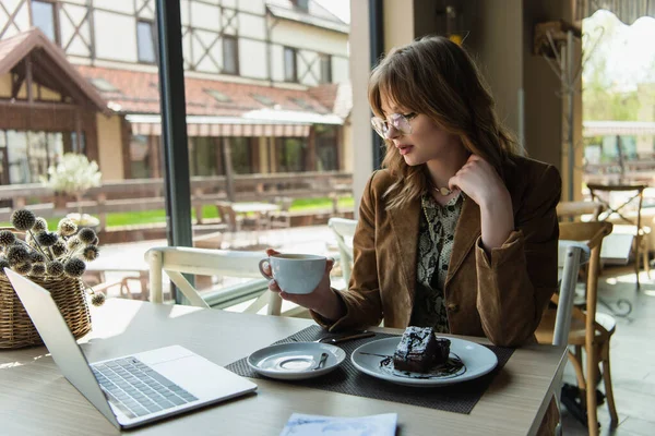 Stylish woman with cup sitting near chocolate dessert and laptop in cafe — Stock Photo