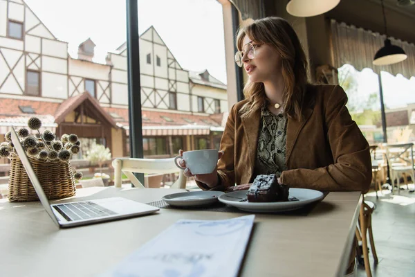 Mulher na moda segurando copo perto de sobremesa, laptop e menu borrado no café — Fotografia de Stock