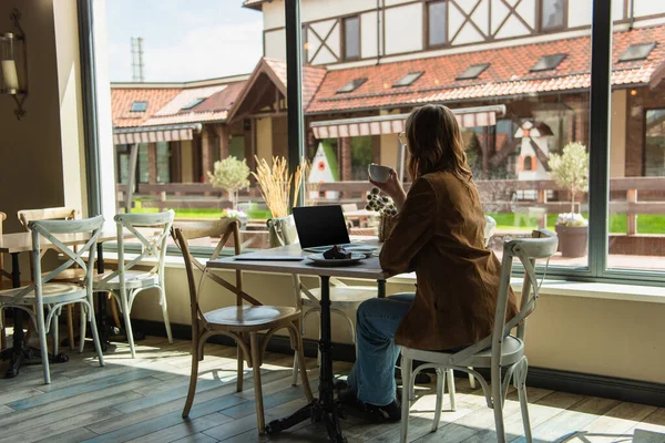 Femme élégante tenant tasse près de délicieux dessert et ordinateur portable dans le café — Photo de stock