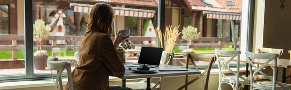 Young woman with cup looking at window near cake and laptop in cafe, banner — Stock Photo