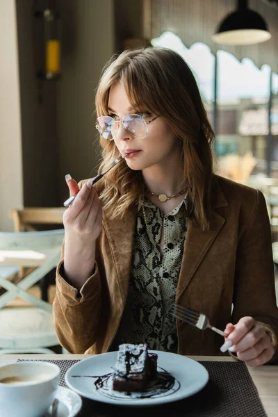 Donna alla moda in occhiali da vista mangiare dessert vicino caffè sfocato in un caffè — Foto stock