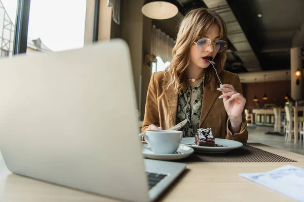 Freelancer che mangia il dessert vicino a caffè e computer portatile sfocato in caffè — Foto stock