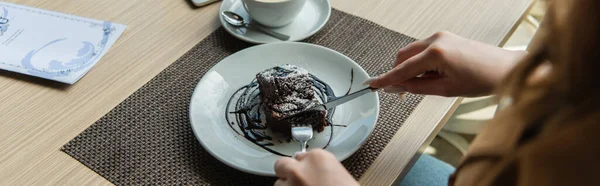 Cropped view of woman cutting dessert near cup and menu in cafe, banner — Stock Photo