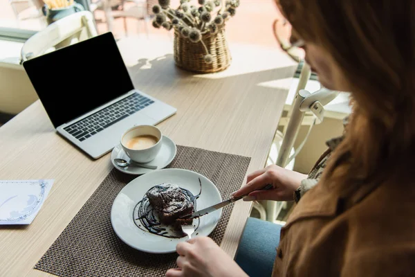 Blurred woman cutting dessert near coffee cup and laptop with blank screen in cafe — Stock Photo