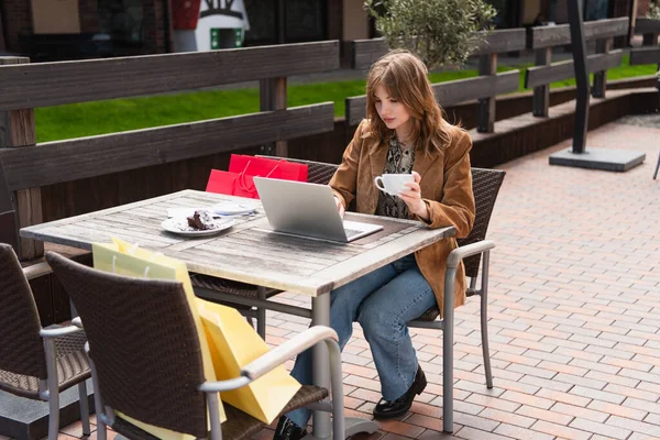 Elegante blogger sosteniendo la taza y usando el portátil cerca de las bolsas de compras en la terraza de la cafetería - foto de stock