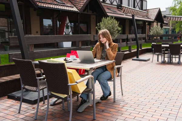 Femme utilisant un ordinateur portable près de tasse, dessert et sacs à provisions sur la terrasse — Photo de stock