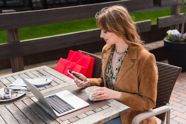 Vista lateral del freelancer con estilo utilizando el teléfono inteligente cerca de café, ordenador portátil y bolsas de compras en la terraza - foto de stock