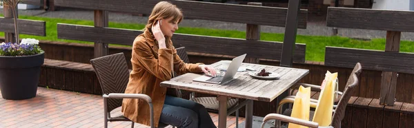 Vista lateral del freelancer en auriculares con portátil cerca de la taza y bolsas de compras en la terraza de la cafetería, pancarta - foto de stock