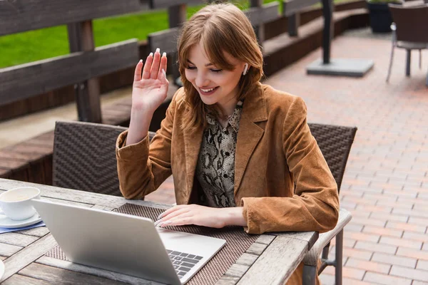 Smiling woman having video call on laptop near cup on terrace of cafe — Stock Photo