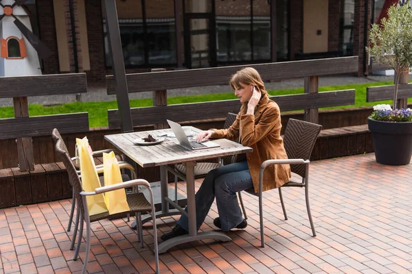 Side view of stylish woman in earphone using laptop near coffee and shopping bags on terrace of cafe — Stock Photo