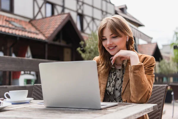 Freelance sorridente in auricolare usando il computer portatile vicino a caffè su terrazza — Foto stock