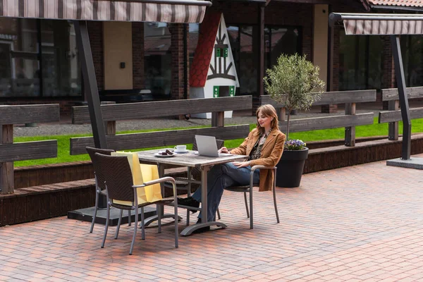 Freelancer de moda con portátil cerca de café, postre y bolsas de compras en la terraza de la cafetería - foto de stock